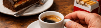 Hand greift nach einer Espressotasse auf einem Holztisch mit Mehlspeise, Zigarettenpackung und Feuerzeug – Sinnbild für Gewohnheiten und Nervensystem-Regulation.